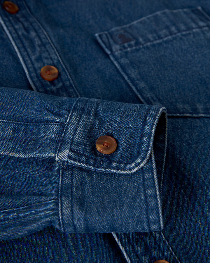 Close-up of a denim shirt sleeve from The Cartagena Denim Shirt - Indigo by Dandy Del Mar, showcasing its collared button-up design. The visible cuff is buttoned with a brown button, highlighting the medium blue denim fabric and white stitching detail.