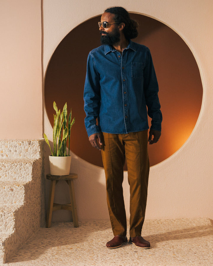 A person stands indoors near stairs, sporting The Cartagena Denim Shirt - Indigo by Dandy Del Mar with a button front placket, brown pants, and sunglasses. A potted plant is placed on a stool next to them.