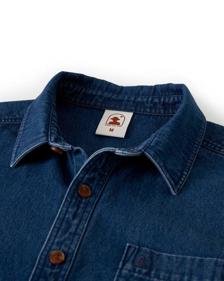 Close-up image of the Cartagena Denim Shirt from Dandy Del Mar, an indigo tailored fit denim collared button-up with brown buttons and a visible collar. The shirt features a brand label indicating size M.