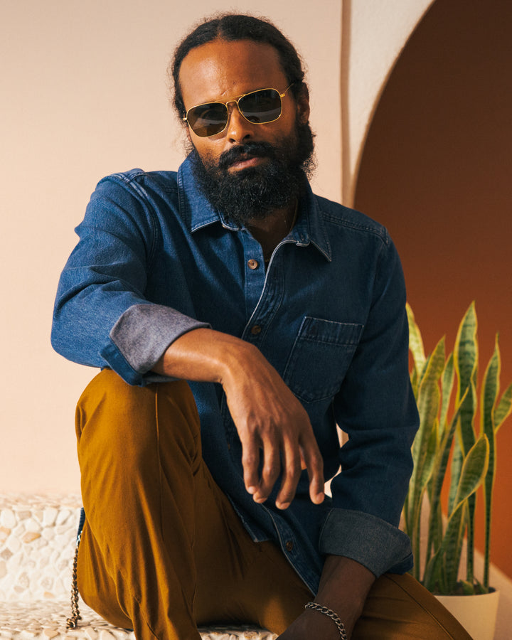 A person with sunglasses, a beard, and hair tied back, wearing The Cartagena Denim Shirt in Indigo by Dandy Del Mar paired with mustard trousers, is sitting in front of a plant and archway background.