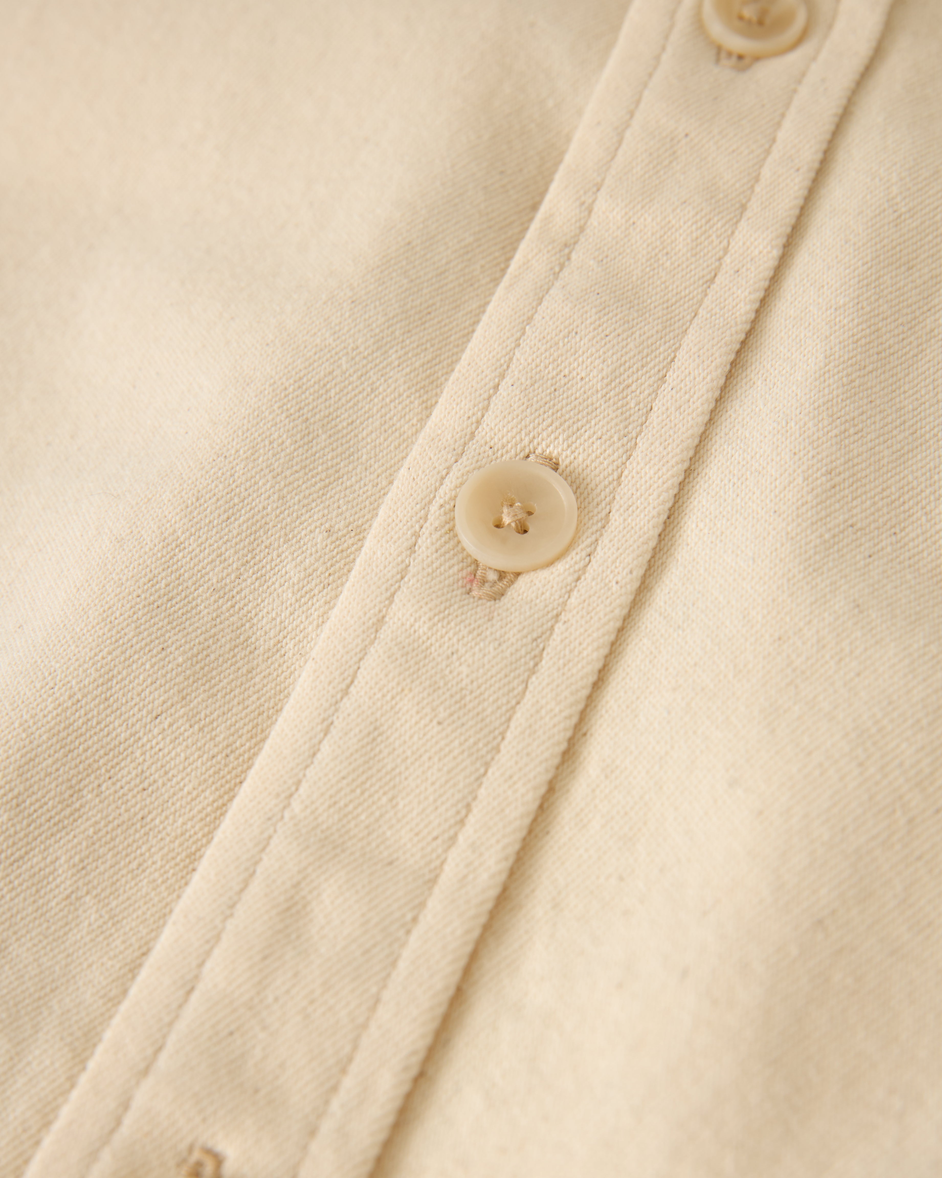 Close-up of The Cartagena Denim Shirt - Scallop by Dandy Del Mar, featuring a beige fabric with light brown buttons.
