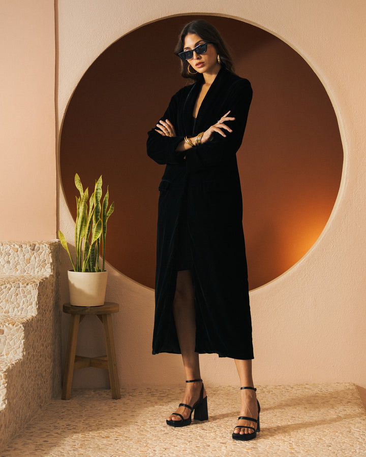 A woman stands with arms crossed in a black dress and sunglasses in front of a circular, orange backdrop. Draped over her shoulders is the luxurious Catania Velour Coat - Onyx from Dandy Del Mar, featuring a shawl collar. To her side, a green potted plant sits gracefully on a wooden stool.