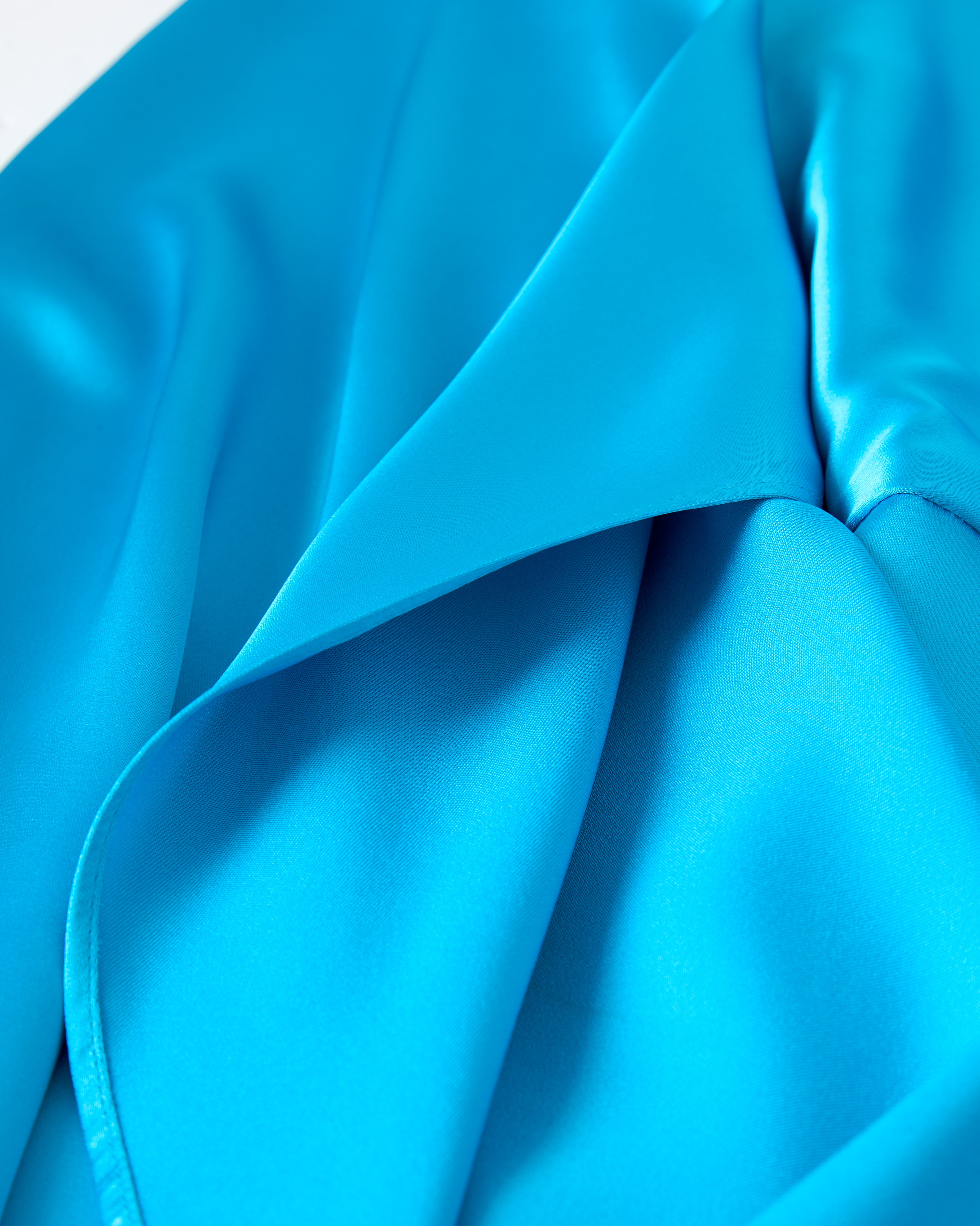 Close-up of The Catania Satin Angel Sleeve Dress - Lago by Dandy Del Mar, showcasing its vibrant blue satin fabric with smooth folds and a glossy finish.