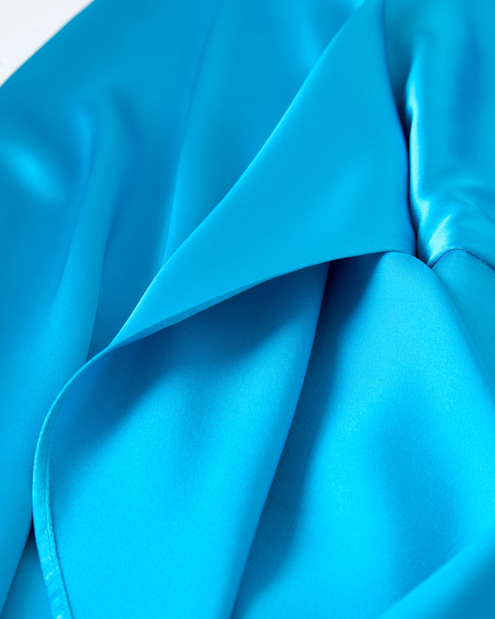 Close-up of The Catania Satin Angel Sleeve Dress - Lago by Dandy Del Mar, showcasing its vibrant blue satin fabric with smooth folds and a glossy finish.