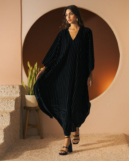 A woman in The Marsala Kaftan - Onyx by Dandy Del Mar stands indoors, posing near a potted plant and a circular wall feature. She wears black sandals and gazes to her left.