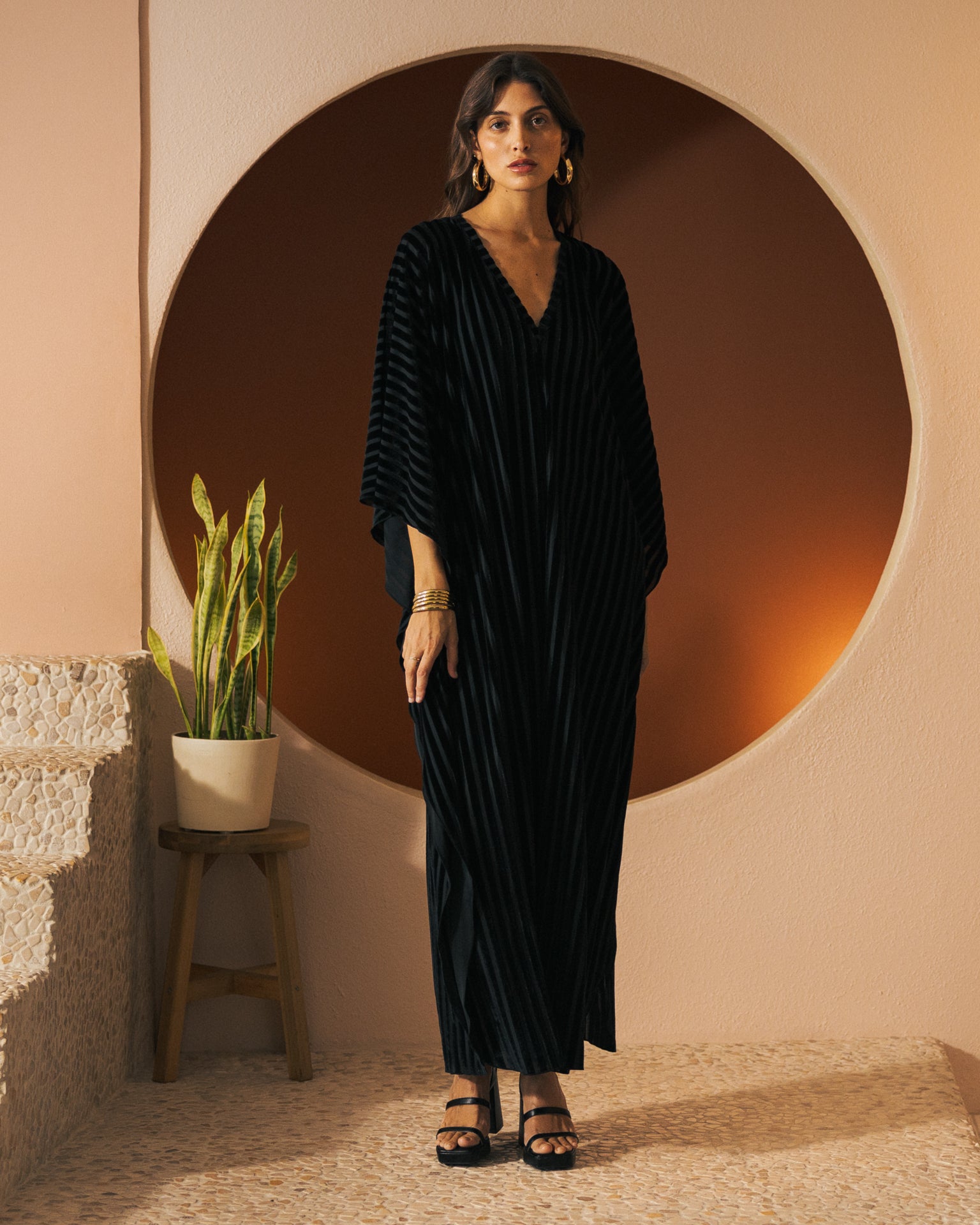 A woman wearing The Marsala Kaftan - Onyx by Dandy Del Mar stands indoors in front of a large circular wall design. She accessorizes with gold hoop earrings and black sandals, while a potted plant rests on a stool beside her.