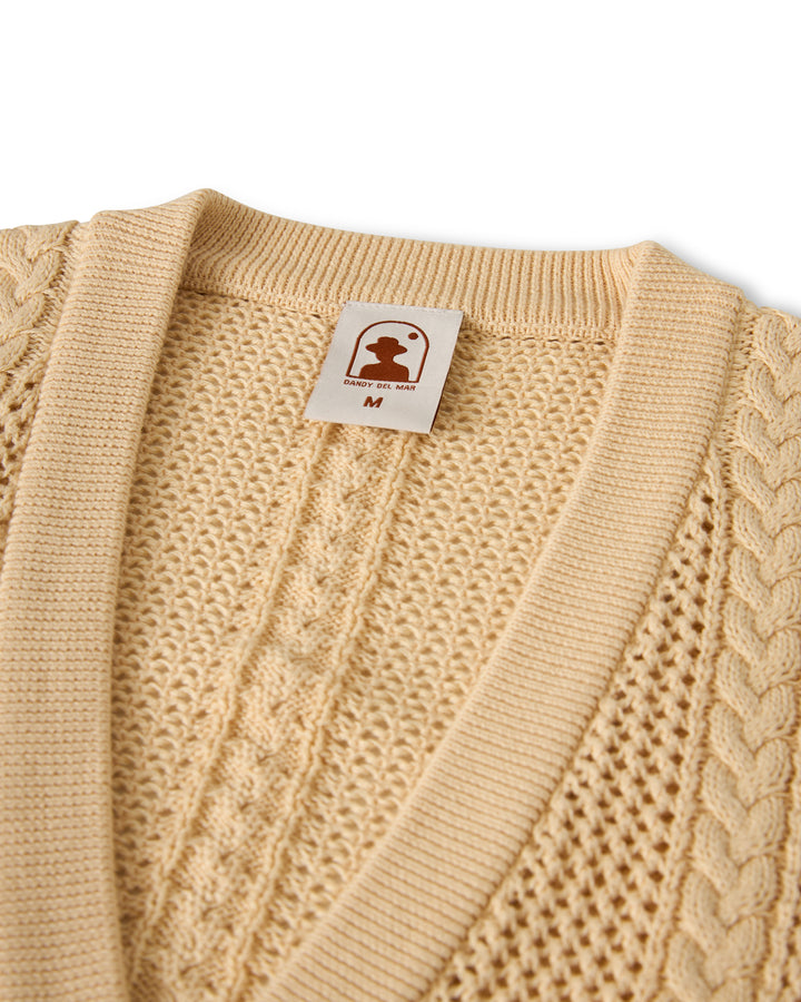 Close-up of The Crete Crochet Cardigan - Sabbia by Dandy Del Mar, featuring a beige cable-knit pattern and a visible "M" clothing tag.