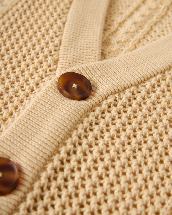 A detailed view of The Crete Crochet Cardigan in Sabbia by Dandy Del Mar, featuring brown buttons and a textured pattern.