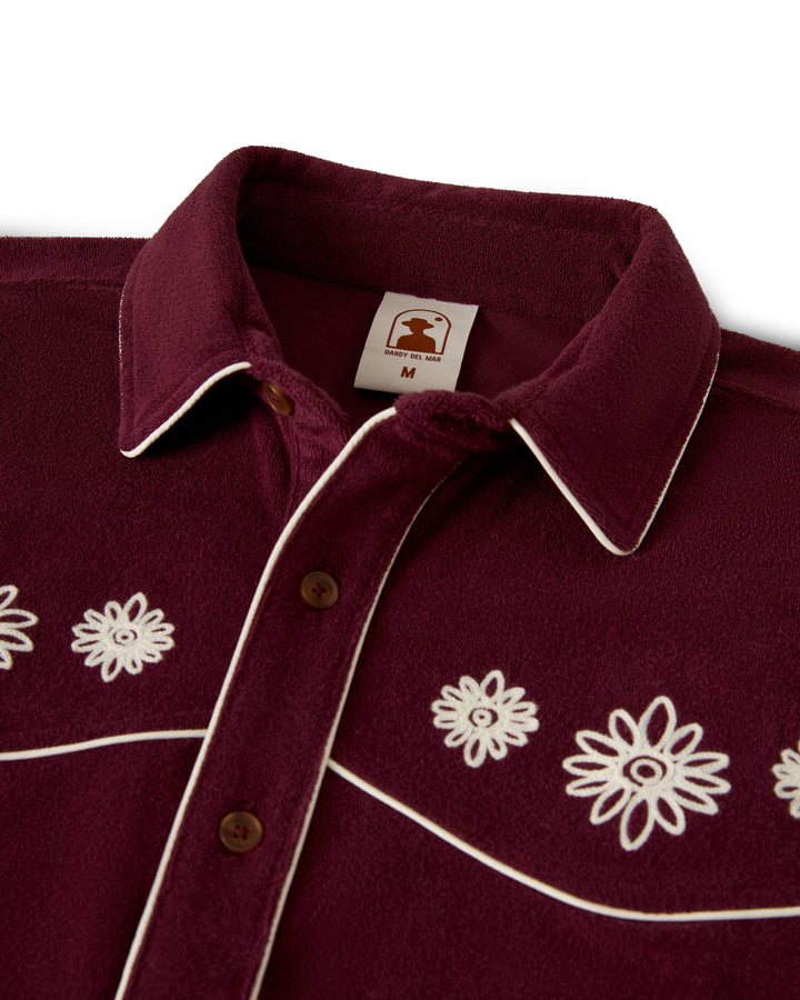 The Gaucho Terry Cloth Shirt in Bordeaux by Dandy Del Mar features white floral embroidery on the front, with trim on the collar and button placket. Brown buttons and a size tag are also visible.