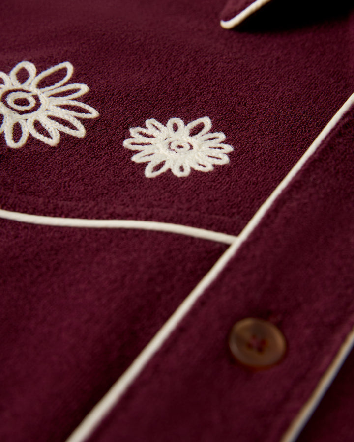 Close-up of The Gaucho Terry Cloth Shirt - Bordeaux by Dandy Del Mar, highlighting maroon fabric with white floral embroidery, brown buttons, and white piping along the edges.