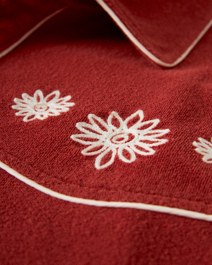 Close-up image of The Gaucho Terry Cloth Shirt - Rouge by Dandy Del Mar, showcasing its red fabric adorned with white floral embroidery and white piping details, reminiscent of an embroidered floral chest on a gaucho shirt.