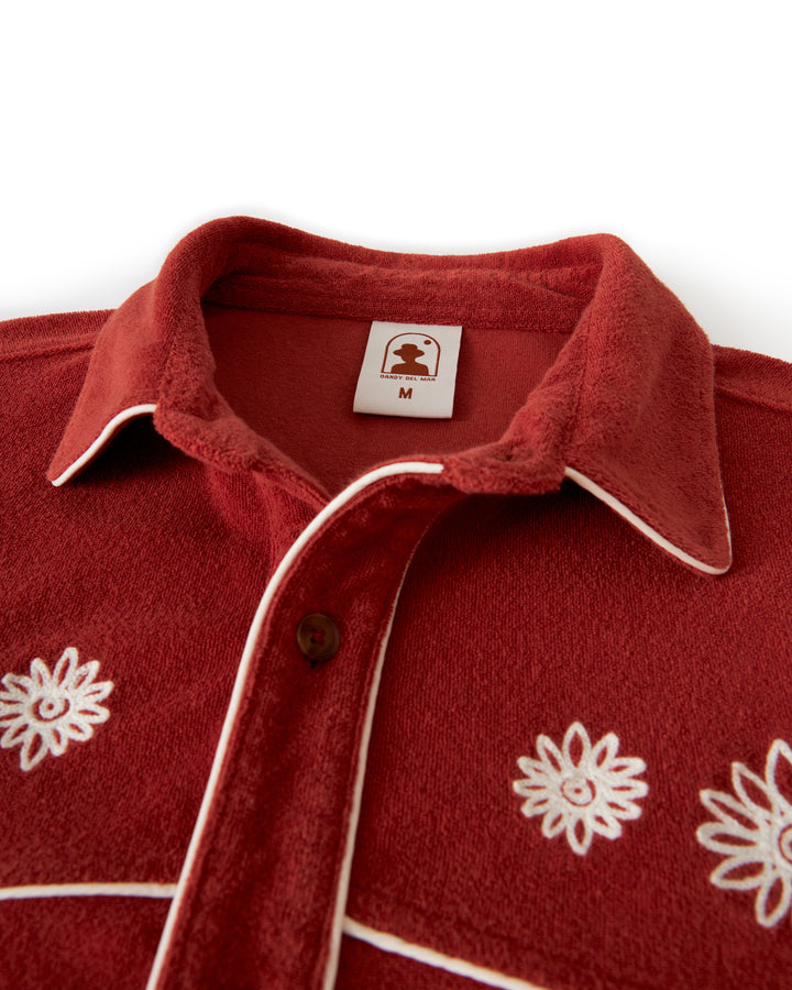 Close-up of a red ultra soft terry cloth shirt from Dandy Del Mar, known as The Gaucho Terry Cloth Shirt - Rouge. It features white embroidered floral chest details and brown buttons. The tag inside the collar displays an icon and the size "M".