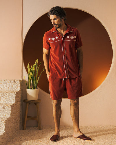 A man stands indoors near a potted plant and circular wall opening, wearing Dandy Del Mar’s The Gaucho Terry Cloth Shorts in Rouge with white embroidery and an elastic waistband.