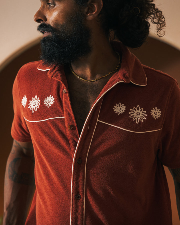 A person with a beard and curly hair is wearing The Gaucho Terry Cloth Short in Rouge, by Dandy Del Mar, featuring white floral embroidery, resembling an Equatorial Cowboy, looking away to the side.