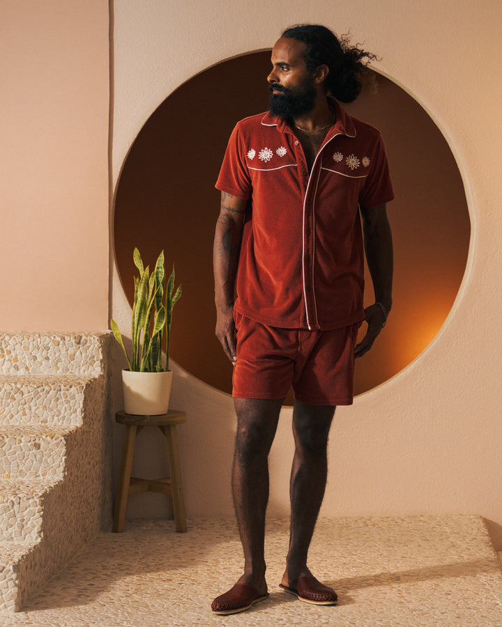 A man with a beard stands in front of a circular wall feature, wearing a red collared shirt and The Gaucho Terry Cloth Short - Rouge by Dandy Del Mar, next to a small potted plant on a stool by tiled steps.