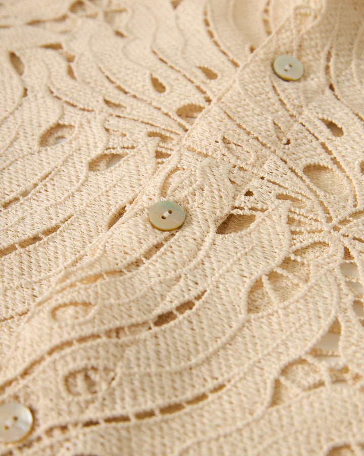 Close-up of The Monstera Shirt - Scallop by Dandy Del Mar, showcasing beige lace fabric with a floral pattern and round buttons.