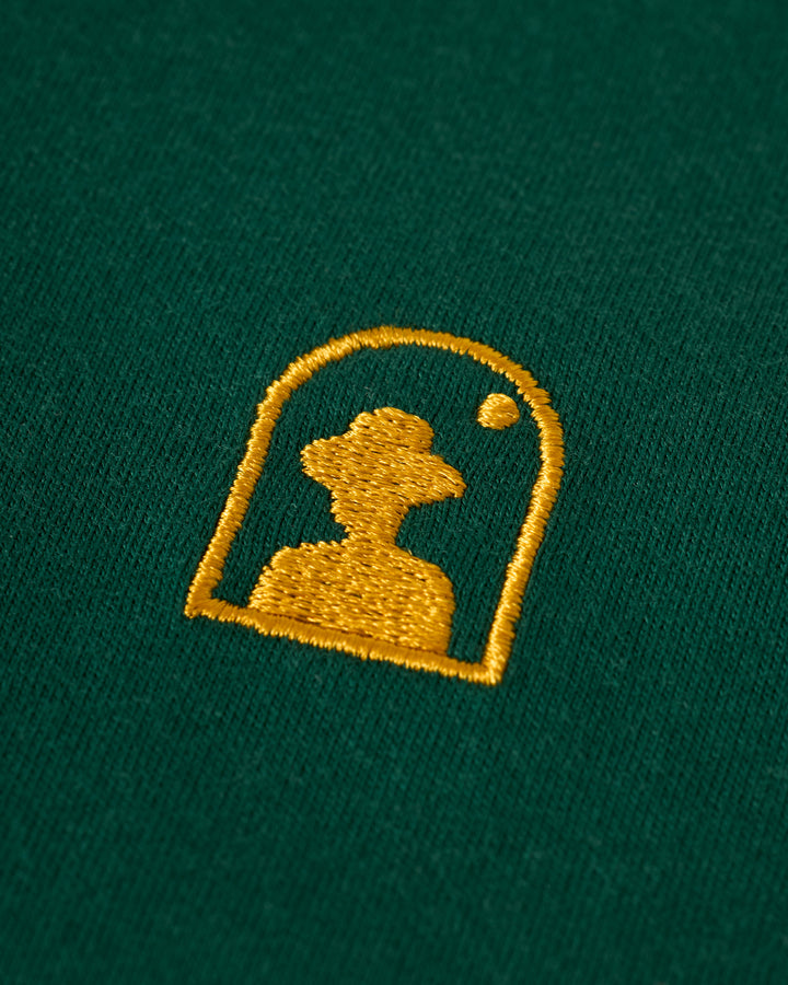 Dandy Del Mar's The Nassau Tee - Albero features a gold embroidered silhouette of a person in a hat on green fabric, framed in an arch shape with a small circle at the top right.