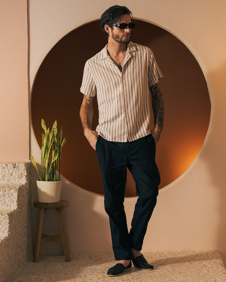 A man with dark hair and sunglasses is wearing The Palma Shirt - Scallop from Dandy Del Mar along with dark pants, standing near a circular wall feature and a potted plant.