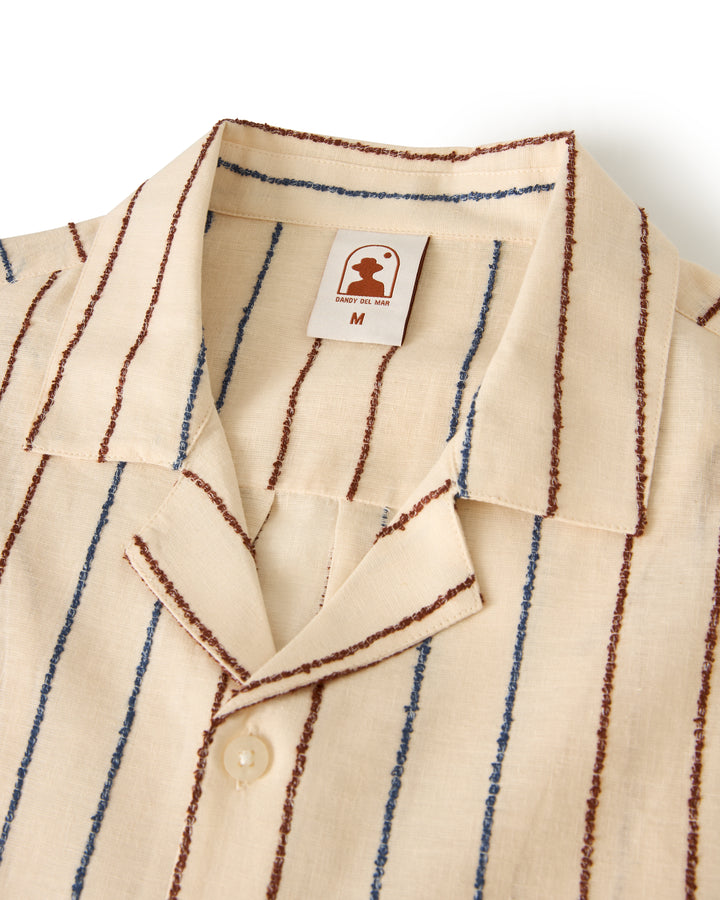 Close-up of The Quarta Shirt in beige with blue and brown vertical stripes and a collar. The label reads "M" with the Dandy Del Mar logo above it.