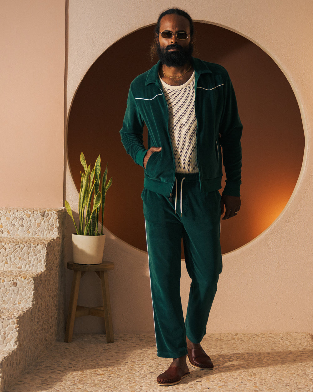 A person with a beard and sunglasses wears The Rio Velour Jacket - Albero by Dandy Del Mar over a white mesh top, standing indoors near a potted plant and a circular wall feature.