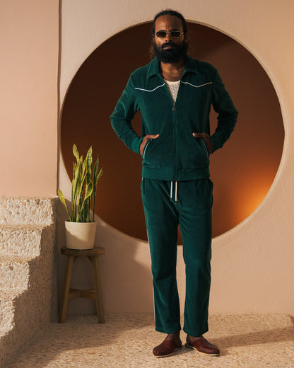 A person with a beard and sunglasses stands in front of a circular backdrop, wearing The Rio Velour Pant - Albero from Dandy Del Mar paired with brown shoes. A potted plant is placed on a stool to the left.