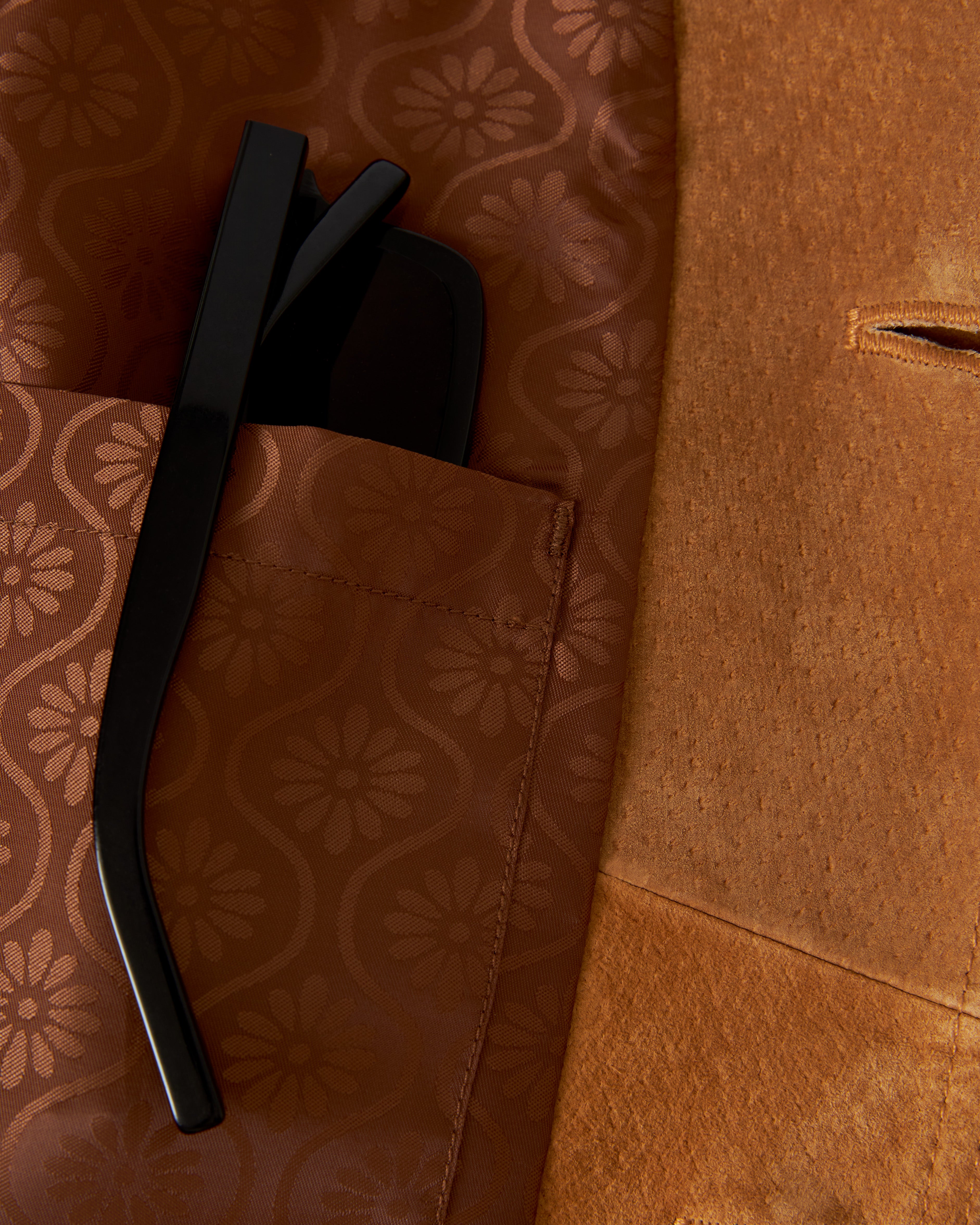 Close-up of a black pair of glasses partially tucked into the patterned brown pocket of The San Juan Suede Leather Jacket - Sable by Dandy Del Mar.