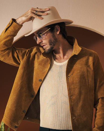 A man wearing The San Juan Suede Leather Jacket - Sable by Dandy Del Mar, a white mesh tank top, and a beige hat touches the brim of his hat while standing in front of an archway.