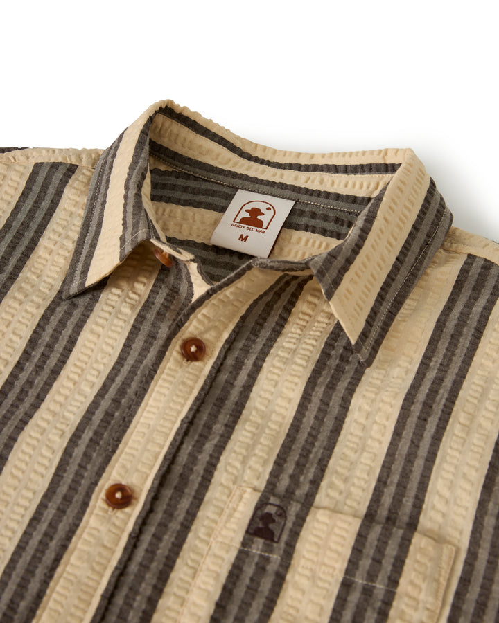 The Santos Shirt - Tierra Weave by Dandy Del Mar is a beige and gray striped button-up with textured fabric, brown buttons, a logo tag inside the collar, and a right-side chest pocket.