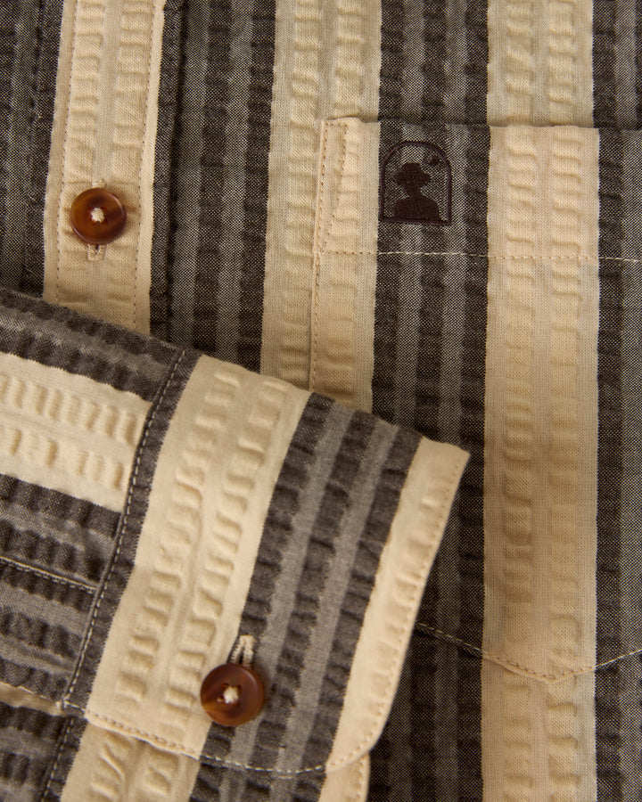 Close-up of The Santos Shirt - Tierra Weave by Dandy Del Mar, showing its striped fabric, chest pocket with brown buttons, and an embroidered silhouette logo above the pocket.