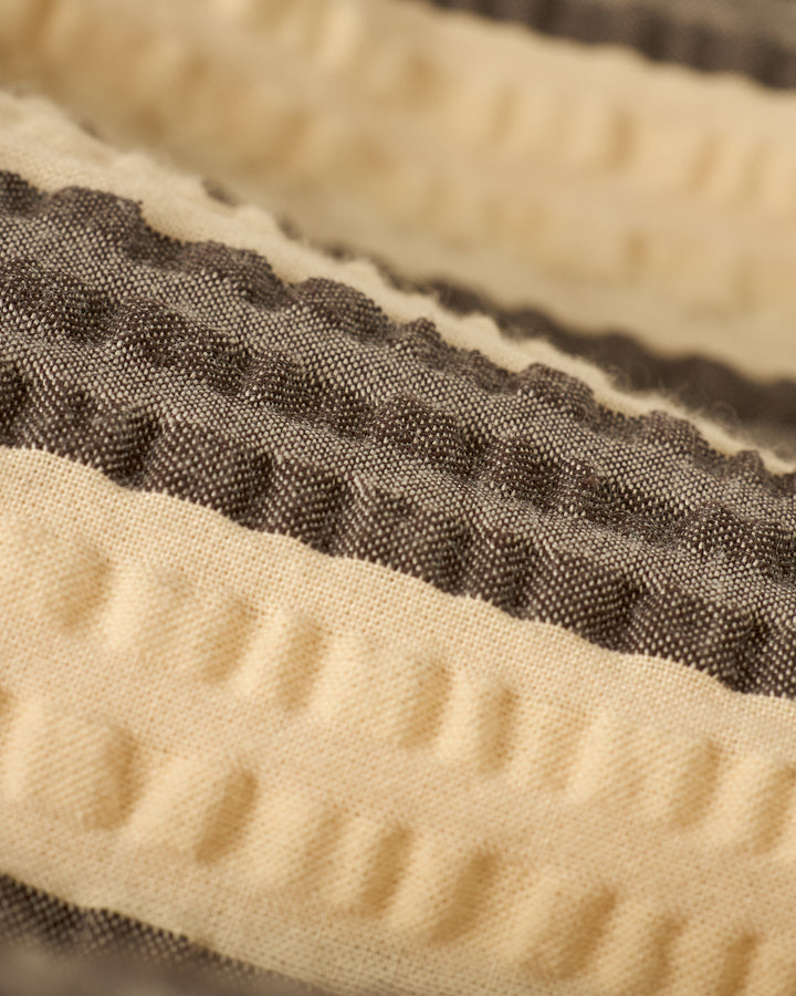 Close-up of The Santos Shirt - Tierra Weave by Dandy Del Mar, showcasing its textured fabric with alternating beige and gray stripes in a wavy pattern.