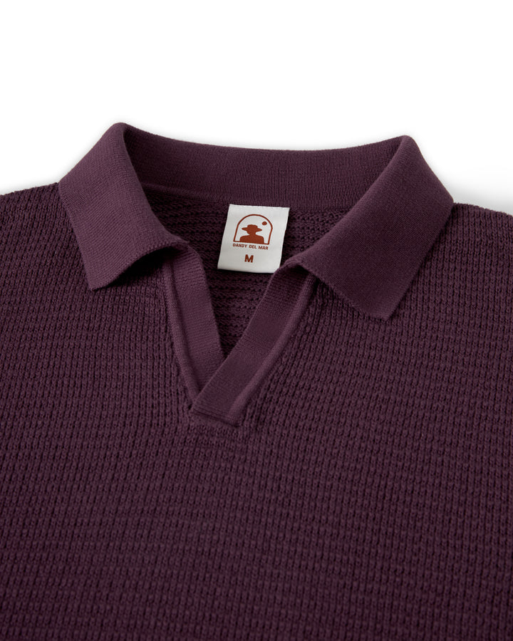 Close-up of The Sebastian Long Sleeve in Amethyst from Dandy Del Mar, showcasing a dark purple hue with amethyst tones, a collar, and an open V-neck. This comfortable shirt is crafted from sweater knit cotton, and includes a small tag inside marked with size "M" and the brand's logo.