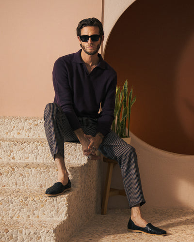 A man in The Sebastian LS Polo Shirt - Amethyst by Dandy Del Mar, checkered pants, black loafers, and sunglasses sits on tiled steps by a potted plant against a neutral-toned backdrop.