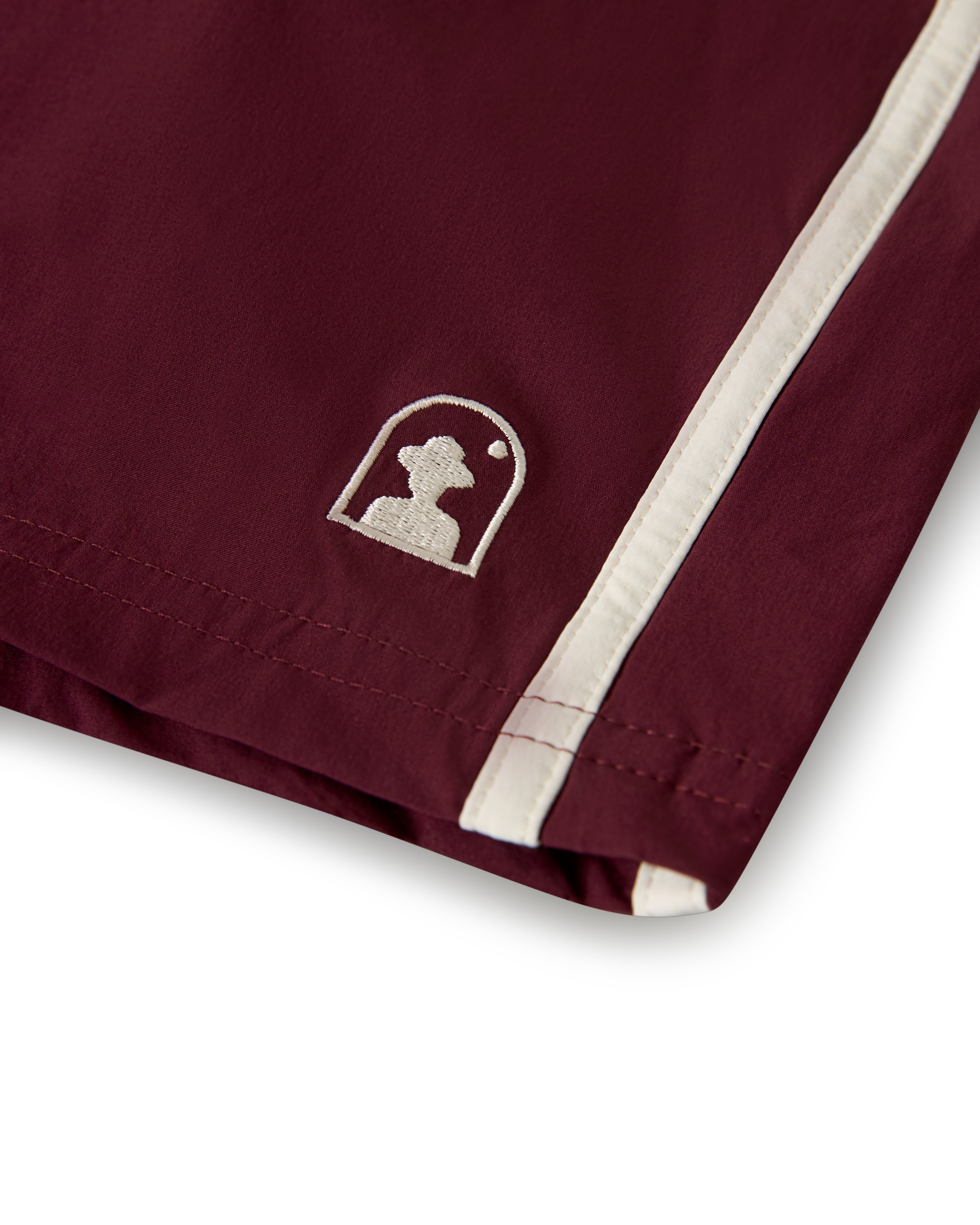 A close-up of The Stirata Swim Short - Bordeaux by Dandy Del Mar shows maroon fabric with a white embroidered logo on the left side and white stripe detailing.