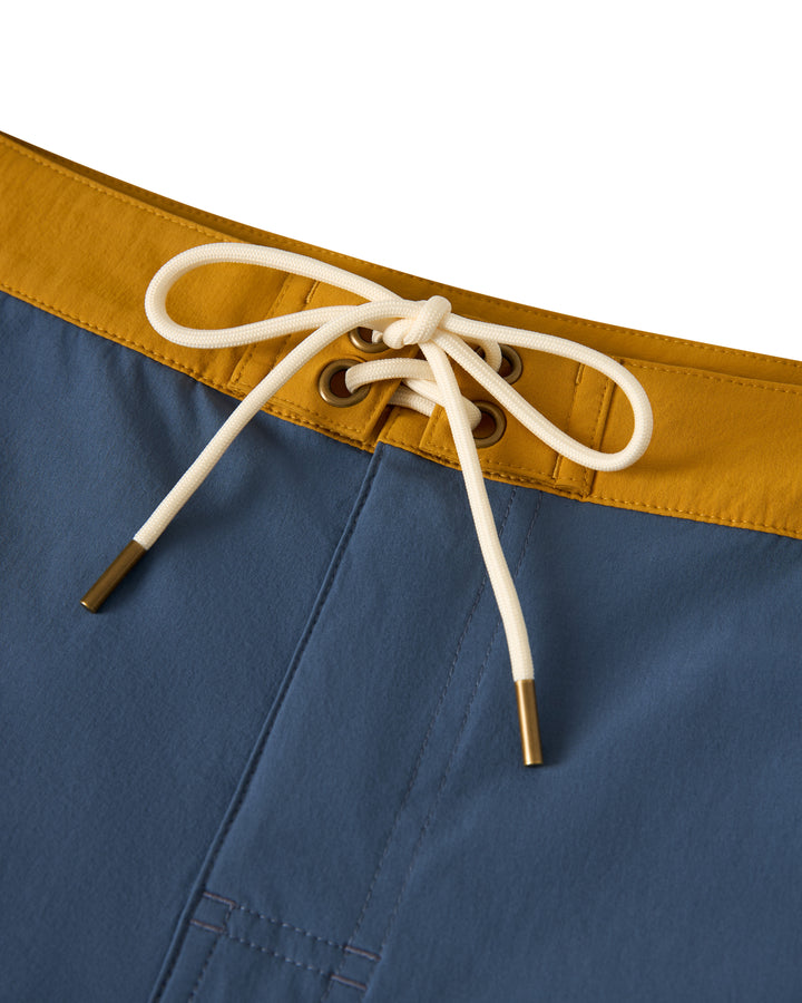 Close-up of The Stirata Swim Short - Moontide by Dandy Del Mar, featuring blue fabric with a yellow waistband and a white drawstring bow.