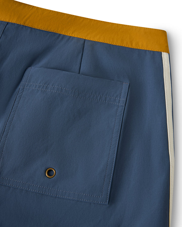 Close-up of The Stirata Swim Short - Moontide by Dandy Del Mar, featuring a blue design with a mustard waistband, white side stripe, and large back pocket with a metal eyelet.