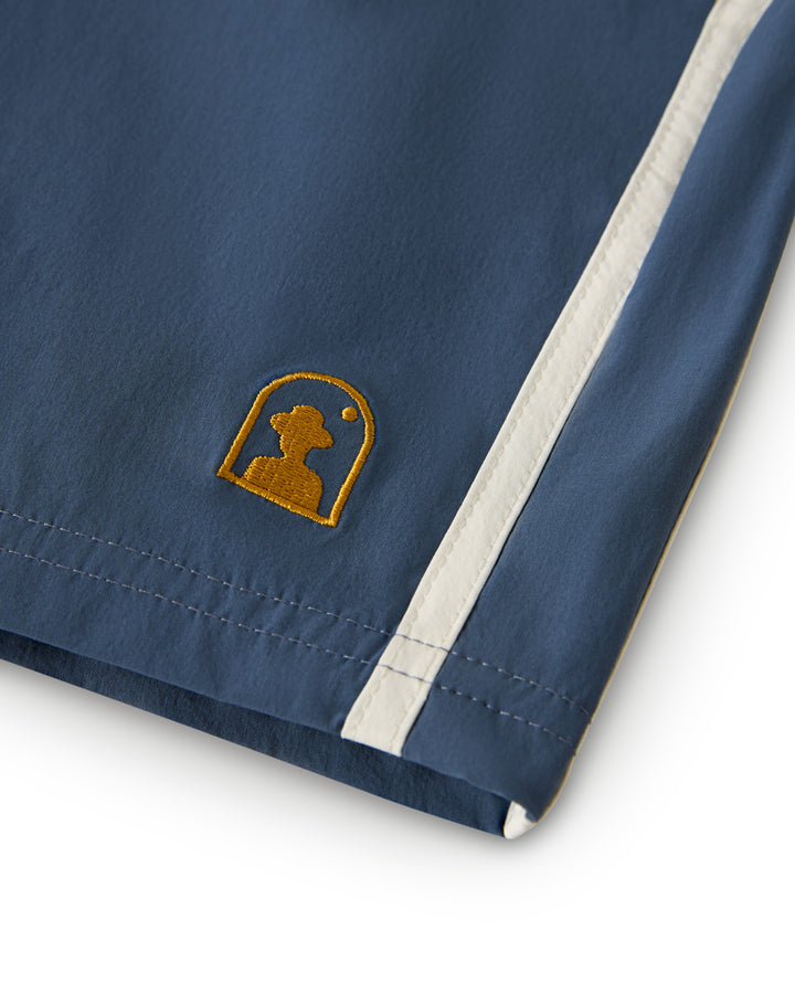 A close-up of the blue Stirata Swim Short by Dandy Del Mar shows a white stripe on the right and a small yellow logo—featuring a stylized profile of a person within an oval.