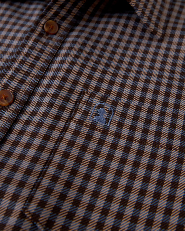 Close-up of the Tresco Shirt - Tierra Houndstooth by Dandy Del Mar, a brown and black checkered, woven collared button-up with wooden buttons and a small embroidered logo of a person in profile on the pocket, exuding casual elegance.