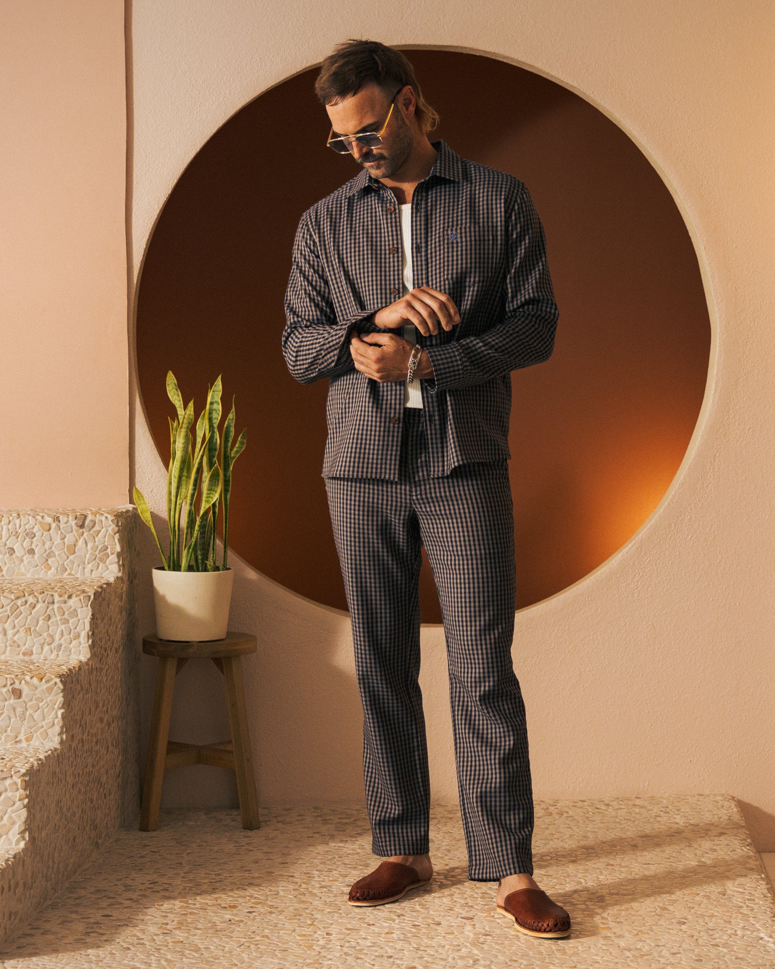 A man in sunglasses and a checkered suit stands next to a potted plant in a modern, minimalist interior, showcasing Dandy Del Mar's latest Tierra Houndstooth pattern from The Tresco Trouser collection.