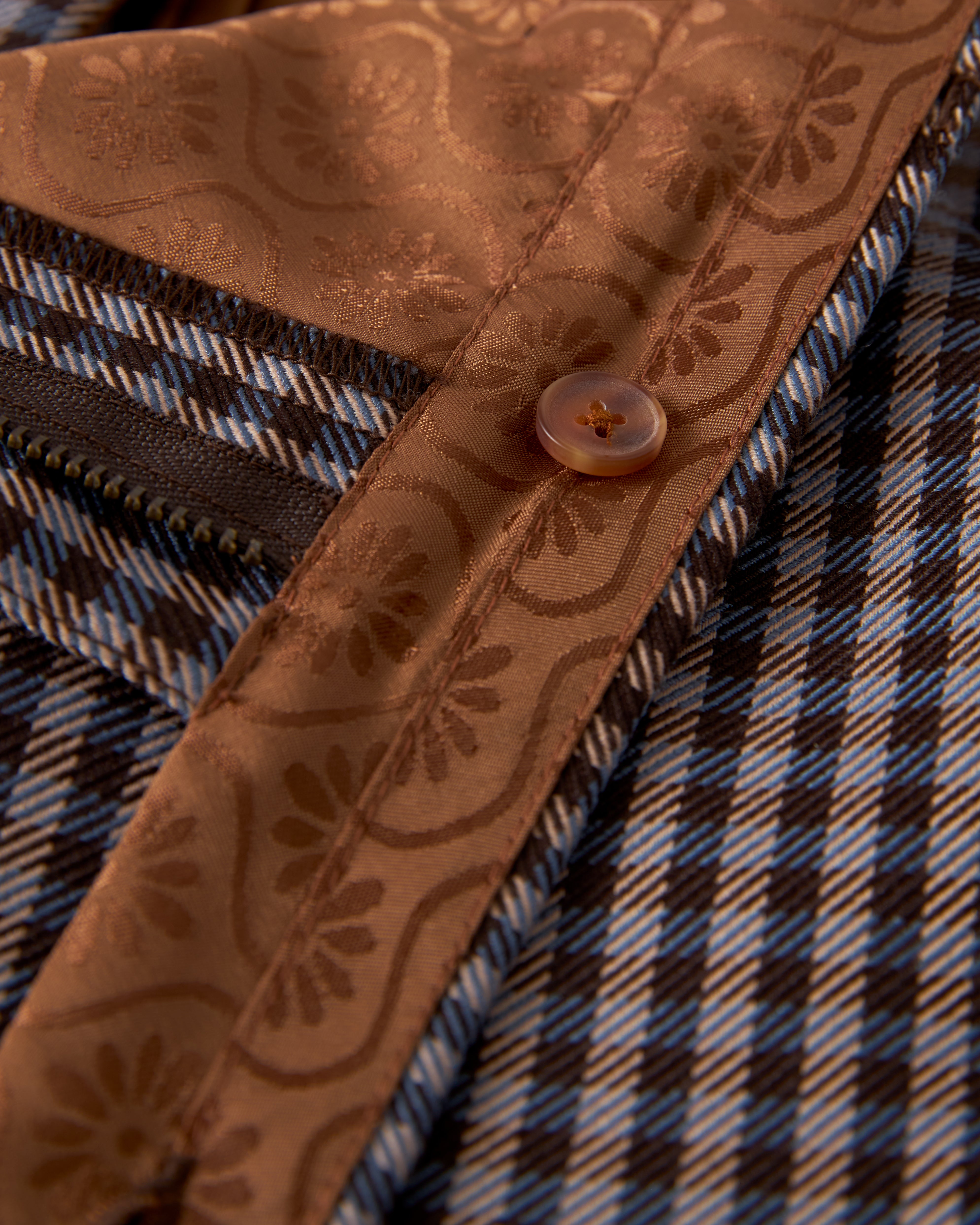 Close-up image of a button on a garment featuring blue and brown plaid fabric with a brown satin trim adorned with floral patterns, evoking the sophistication of The Tresco Trouser - Tierra Houndstooth by Dandy Del Mar.