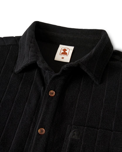 The Tropez Jacquard Shirt - Onyx by Dandy Del Mar boasts a sleek design with a black collar, brown buttons, and a chest pocket. Made from premium terry cloth, it embodies Côte d'Azur elegance. An "M" label sits below the stylized logo inside.