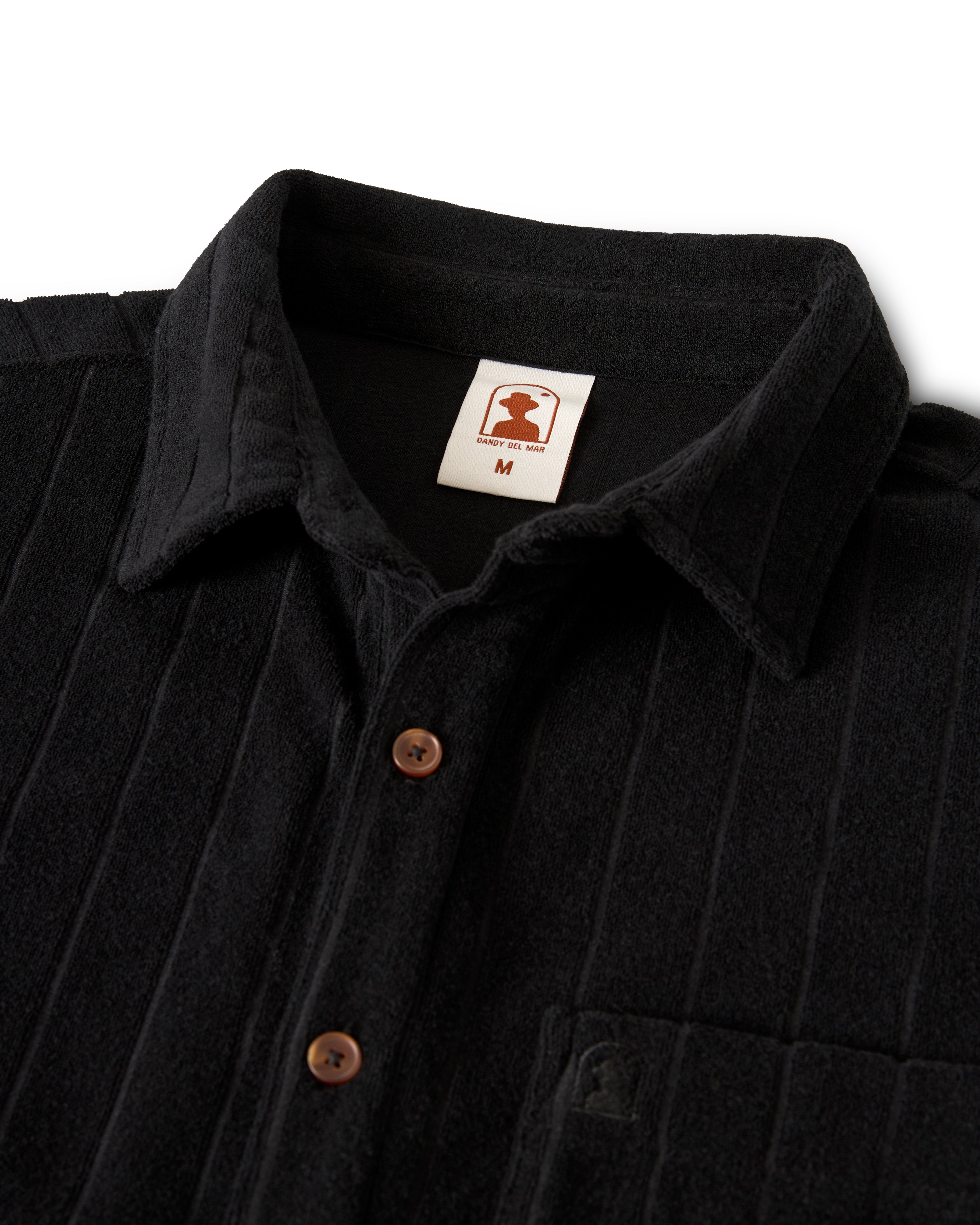 The Tropez Jacquard Shirt - Onyx by Dandy Del Mar boasts a sleek design with a black collar, brown buttons, and a chest pocket. Made from premium terry cloth, it embodies Côte d'Azur elegance. An "M" label sits below the stylized logo inside.