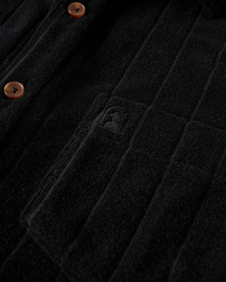 Close-up of The Tropez Terry Cloth Shirt in Onyx by Dandy Del Mar, featuring black textured fabric with vertical seams and brown buttons. A subtle emblem is visible near one patch pocket.