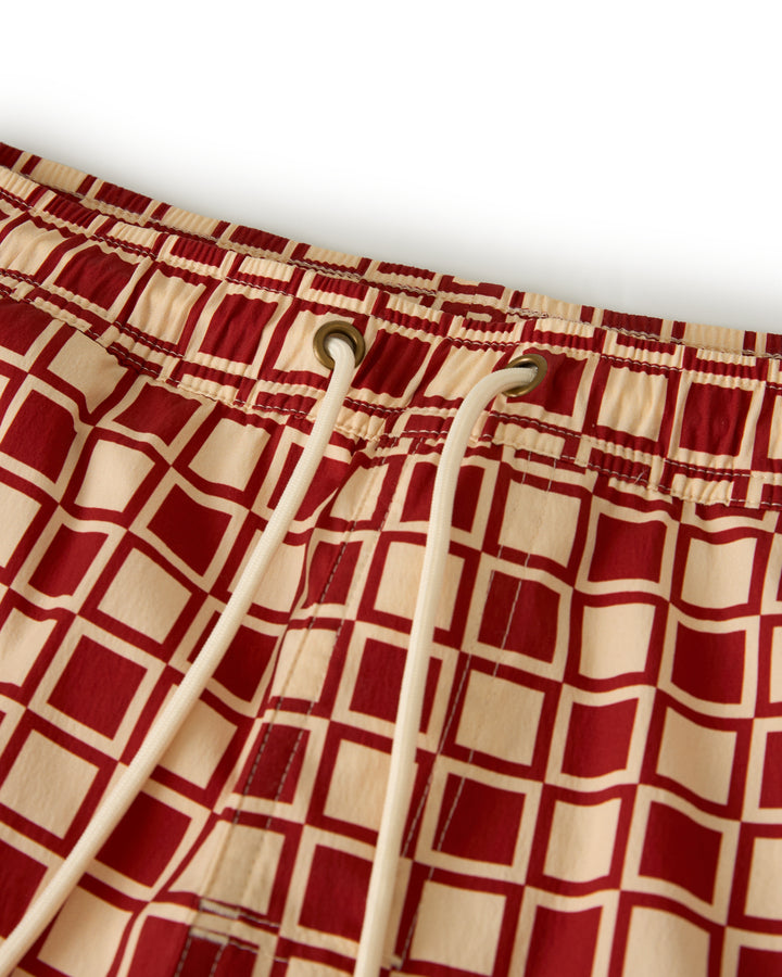 Close-up of The Ventura Volley Short - Rouge by Dandy Del Mar, highlighting the red grid pattern, metal eyelets, and white drawstrings against a beige background.