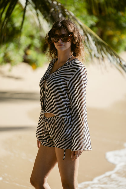 A woman in Dandy Del Mar's Curacao Top - Tierra and sunglasses stands on a sandy beach, with palm trees and gentle waves behind her.