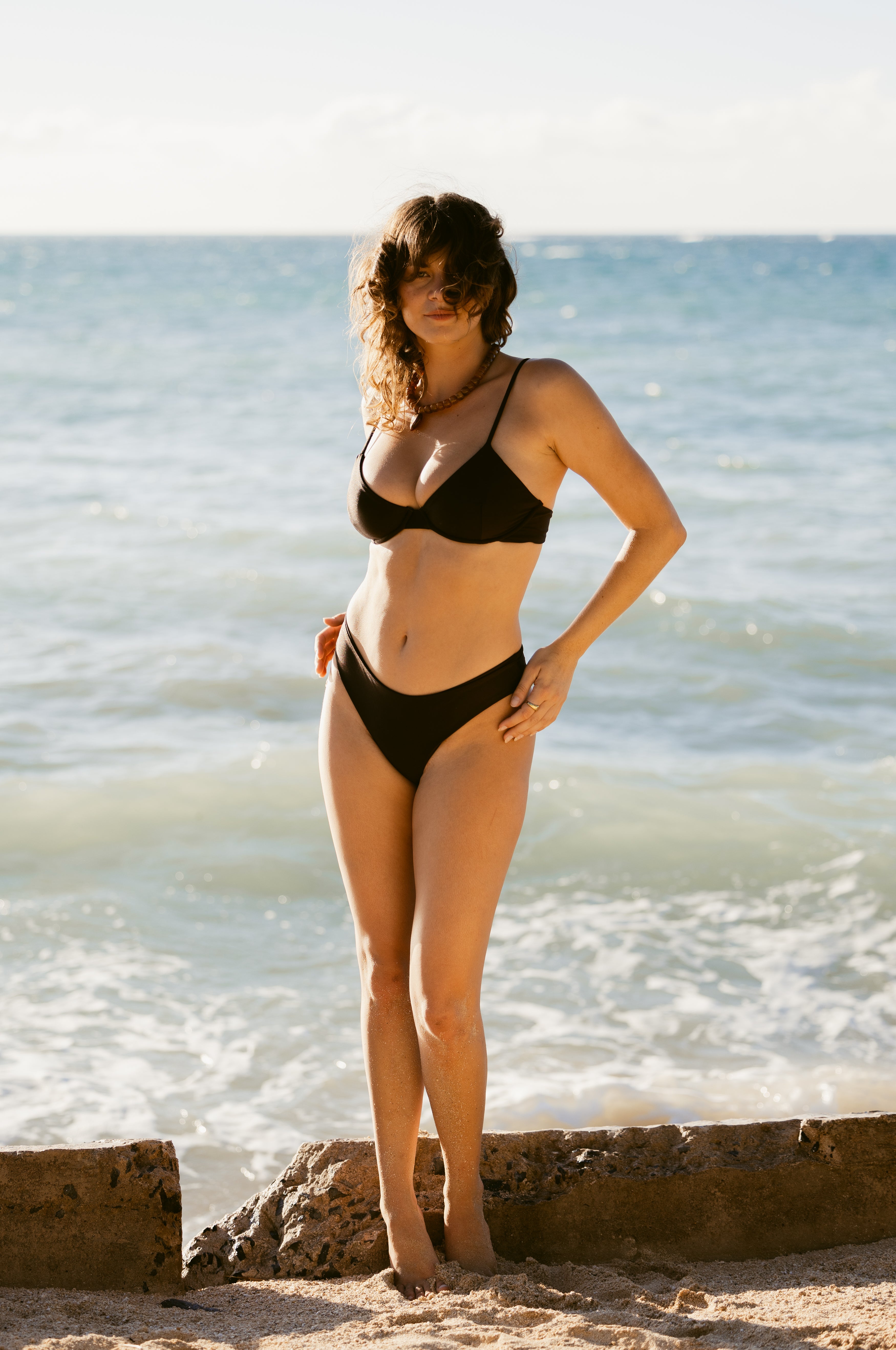 A woman wearing Dandy Del Mar's The Delphi Bottoms - Tierra Shine, featuring moderate coverage, stands on a sandy beach with ocean waves beneath a clear sky.