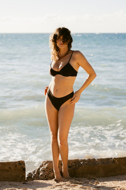 A woman wearing Dandy Del Mar's The Delphi Bottoms - Tierra Shine, featuring moderate coverage, stands on a sandy beach with ocean waves beneath a clear sky.