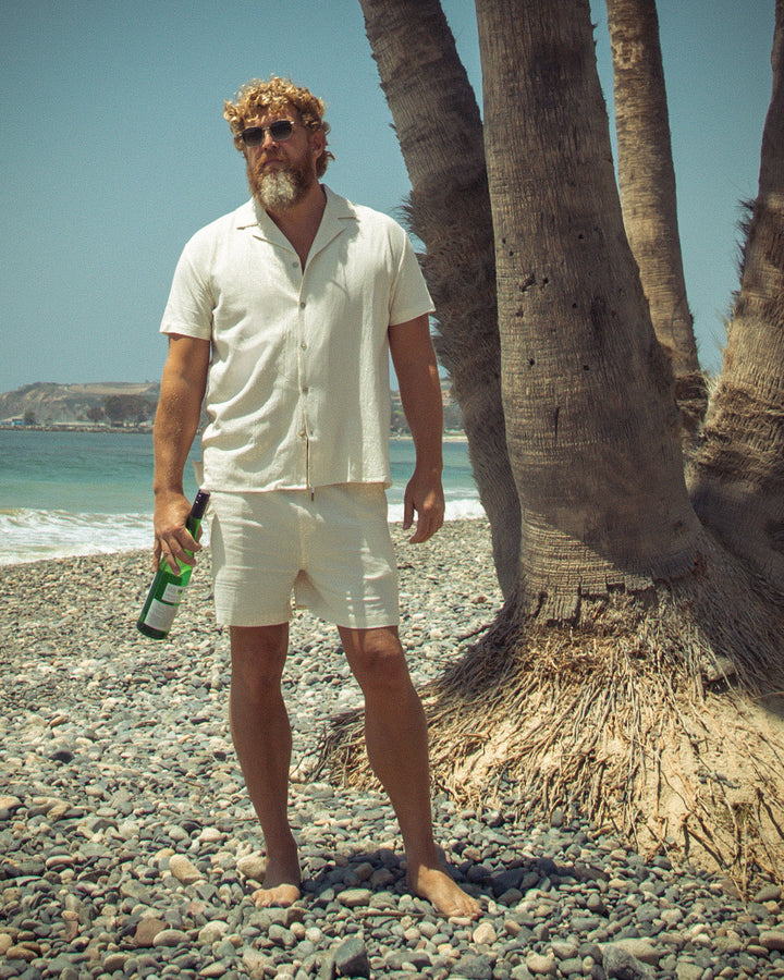 A man standing on a beach next to palm trees wearing Dandy Del Mar's Grenadine Shorts in Vintage Ivory.