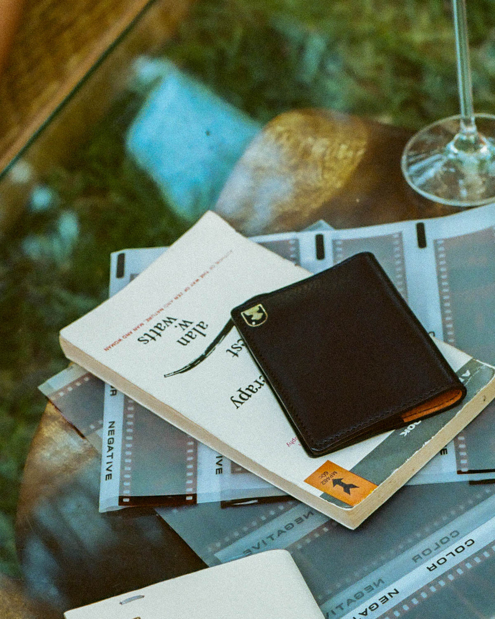 A glass of wine, The Departure Bifold Wallet - Onyx from Dandy Del Mar, and a book on a table.