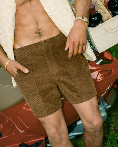 Wearing Dandy Del Mar's The Corsica Corduroy Shorts in Tobacco, a man stands outdoors with an open shirt, holding a crate of wine bottles beside a red scooter.