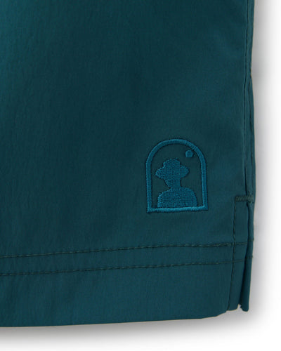 Close-up of teal fabric from The Ventura Volley Shorts - Acqua by Dandy Del Mar, featuring a stitched hem and an embroidered logo of a person's silhouette in profile inside an arch shape.